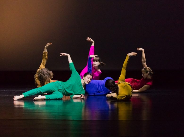 Grupa tancerek w kolorowych strojach leży na podłodze. Ich ciała układają się w kręgu. Wszystkie tancerki podnoszą do góry ręce w identycznym geście. Tło jednolite, ciemne.