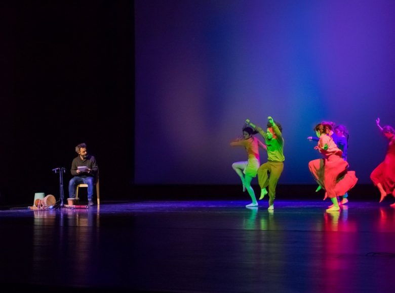 Zdjęcie. W jednej części sceny skaczą tancerki w kolorowych strojach. Po drugiej stronie na krześle siedzi mężczyzna. Obok niego stoi dziecko, w rękach trzyma drewniane pałeczki. Przed chłopcem leży duża, fioletowa piłka.