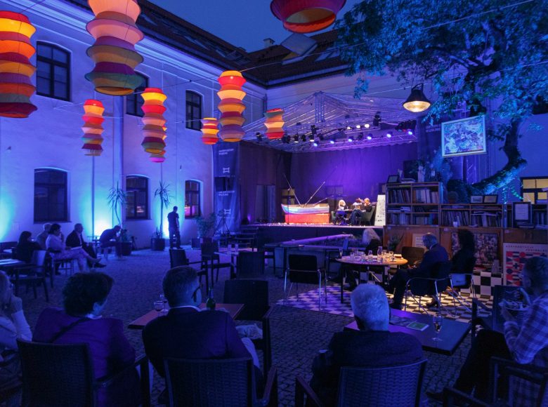 Wirydarz Centrum Kultury w Lublinie. Przestrzeń jest oświetlona kolorowymi lampionami. Na scenie ustawiona jest łódka, wędki i dwa zielone fotele. Przy stolikach siedzą ludzie i oglądają występ.