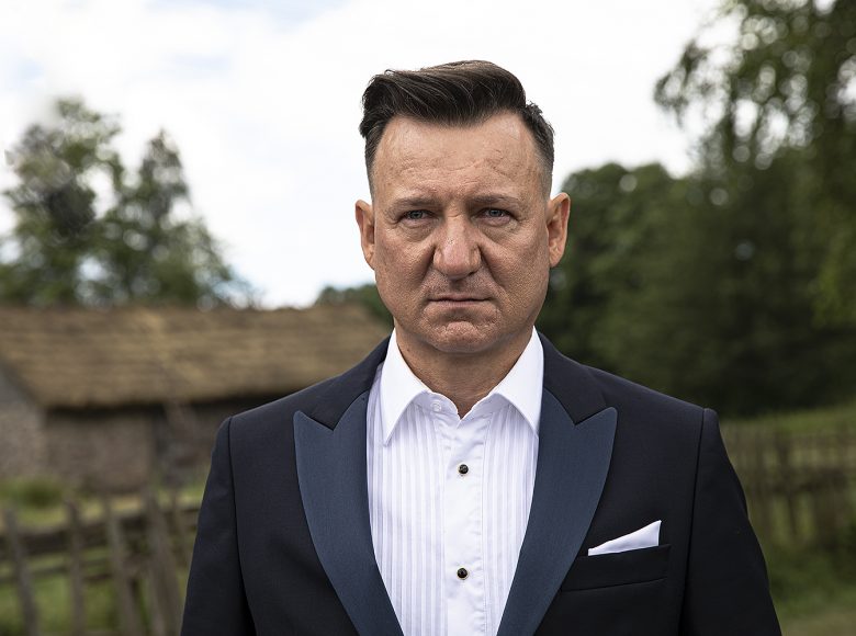 Kadr z filmu. Zbliżenie na elegancko ubranego mężczyznę. W tle rozmazany krajobraz wiejski. Drewniany dom, płot i drzewa.