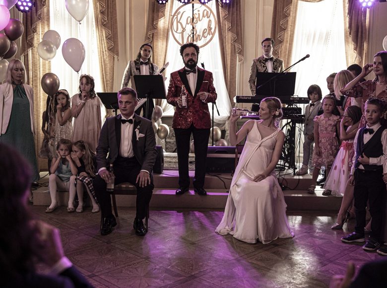 Kadr z filmu. Scena z wesela. Na sali znajduje się orkiestra i goście weselni. Młoda para siedzi obok siebie na krzesłach. Każde z nich trzyma w ręce flaszkę wódki.