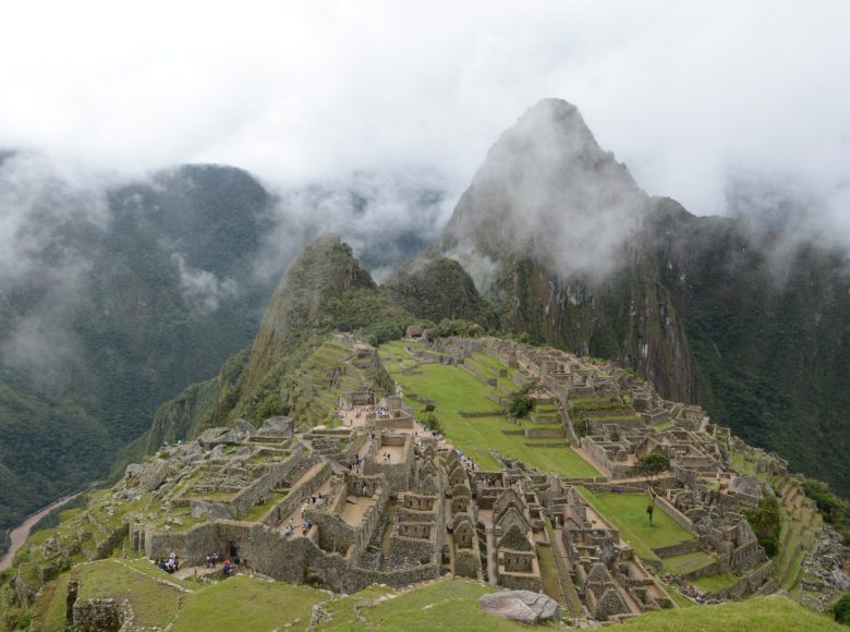 Krajobraz. Widok z góry na starożytne miasto Inków - Machu Picchu. Wzgórze porośnięte zieloną trawą. Na nim kamienne ruiny dawnych budynków, ułożone schodkowo. Widać osoby zwiedzające to miejsce. W tle wysokie góry z wierzchołkami ukrytymi w chmurach.