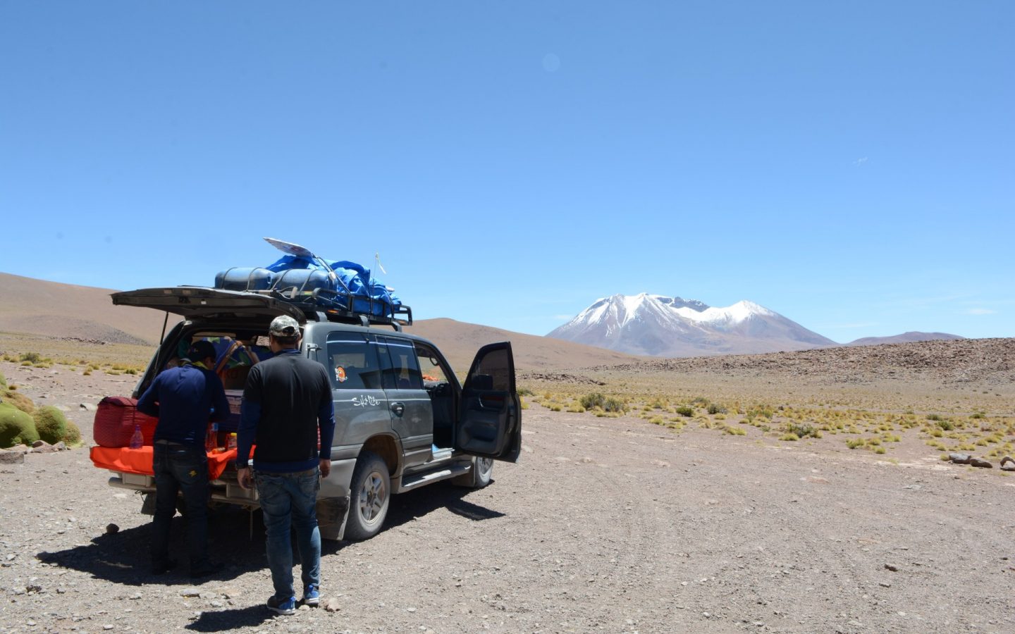 Krajobraz. Na pierwszym planie samochodów terenowy z otwartym bagażnikiem oraz stojącym obok niego dwóch mężczyzn. Samochód jest mocno załadowany, także na dachu przyczepione są bagaże. Auto stoi pośrodku drogi na pustkowiu. Widać żwir, piach i kępki roślinności. W tle wysoka góra z ośnieżonym szczytem. Nad wszystkim czyste, błękitne niebo.