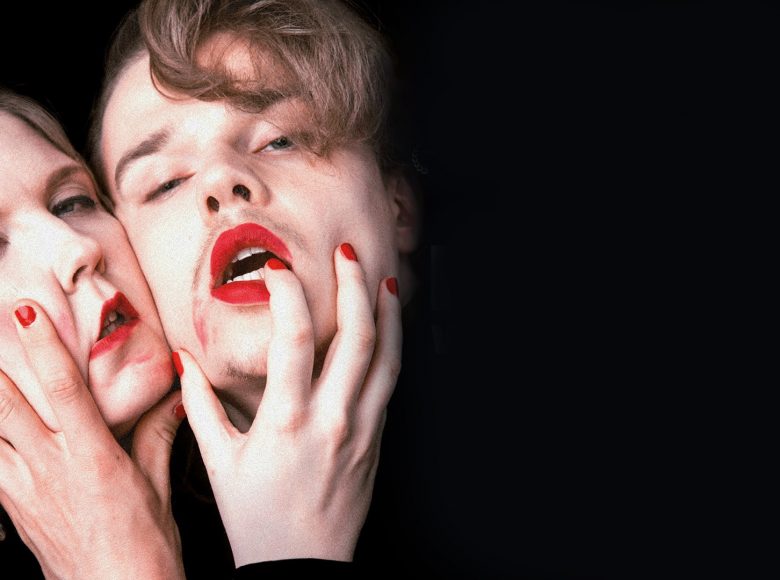 Zdjęcie. Z ciemności wyłaniają się twarze dwóch osób, mężczyzny i kobiety. Są blisko siebie. Usta i paznokcie mają pomalowane na intensywny, czerwony kolor.