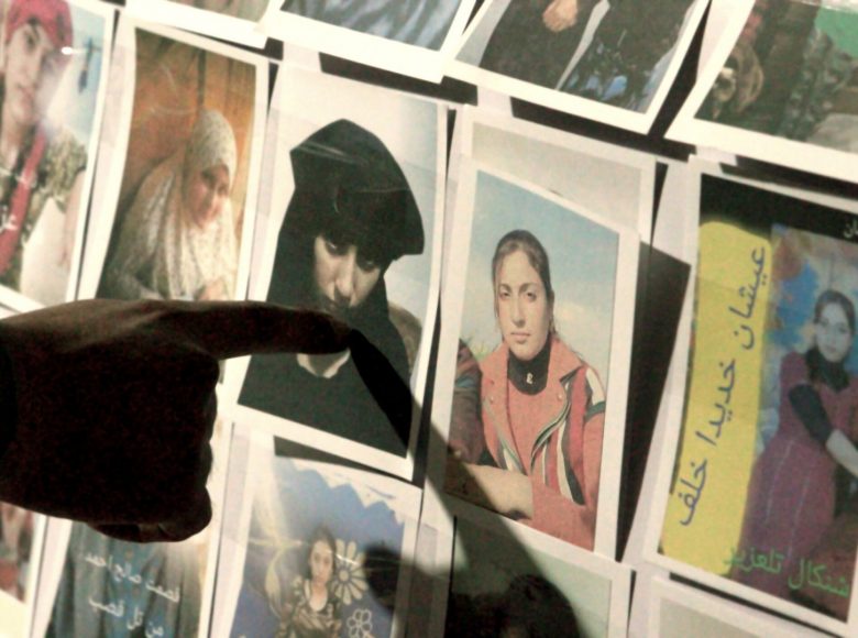 Na ścianie przyczepione wiele zdjęć kobiet w różnym wieku i różnych strojach. Dłoń wskazuje jedno ze zdjęć przedstawiające młodą kobietę w czarnym stroju i nakryciu głowy.