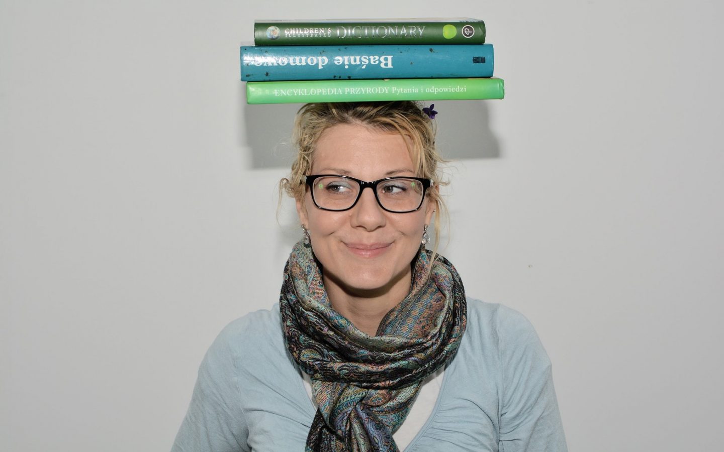 Zdjęcie portretowe uśmiechniętej kobiety w okularach z trzema książkami na głowie.
