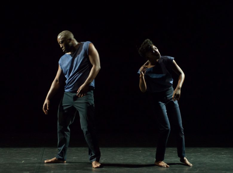Na ciemnej scenie stoją obok siebie czarnoskórzy mężczyzna i kobieta. Każdy z nich jest w innej tanecznej pozie. Mężczyzna lekko pochyla się w bok i patrzy w dół a kobieta podnosi rękę do twarzy. Są boso, w luźnym stroju.