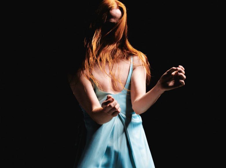 Na czarnym tle stoi dziewczyna w jasnej, niebieskiej sukience. Ma długie, rude włosy zasłaniające jej twarz.