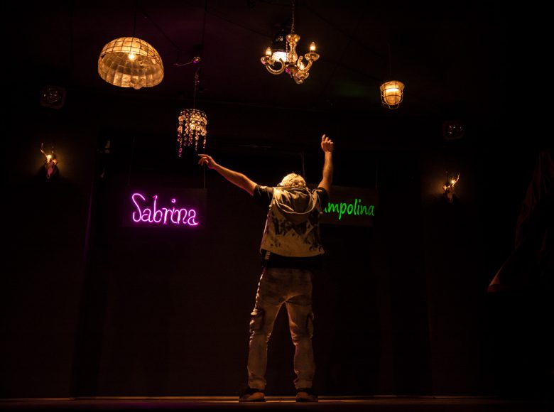 Scena ze spektaklu. Mężczyzna stoi na scenie odwrócony plecami, ręce unosi do góry. Pomieszczenie oświetlają neony i kila różnych lamp.