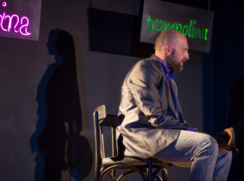 Scena ze spektaklu. Mężczyzna w garniturze siedzi na krześle. Za nim na ścianie świecą dwa neony z napisami trampolina i Sabrina.