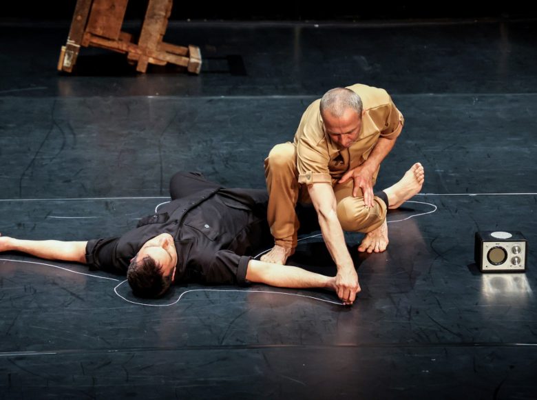 Scena ze spektaklu teatru tańca. Mężczyzna w ciemnym ubraniu leży na podłodze. Mężczyzna w jasnym ubraniu rysuje zarys leżącego kredą.