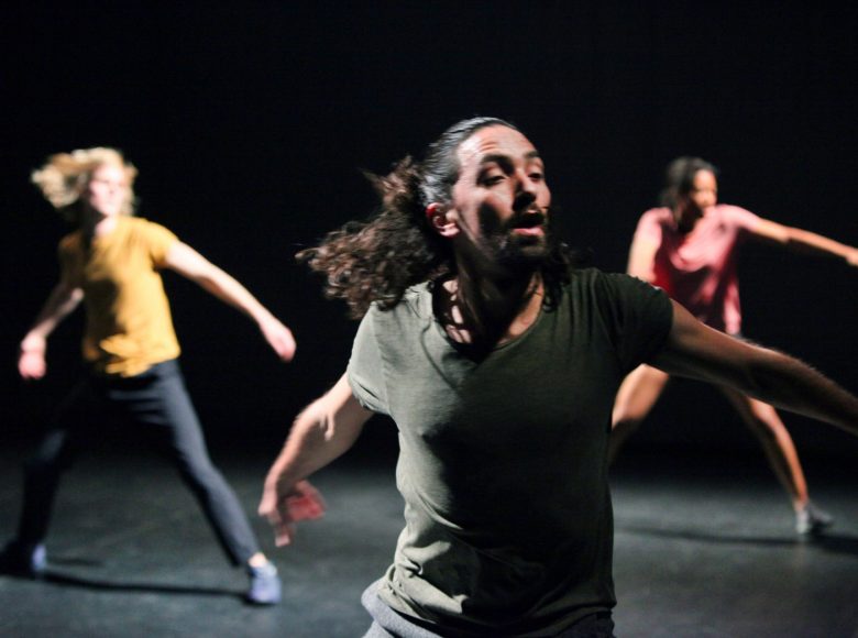 Na czarnej scenie tancerz i dwie tancerki tańca współczesnego w dynamicznych pozach.