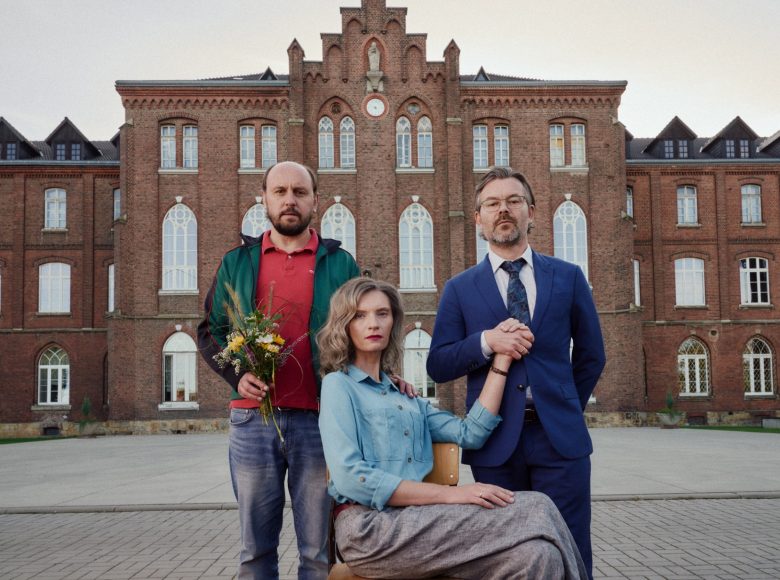 Trójka osób przed budynkiem z cegły. Kobieta centralnie, siedzi na krześle. Po obu jej bokach stoją mężczyźni. Jeden trzyma kobietę za rękę. Drugi trzyma bukiet polnych kwiatów.