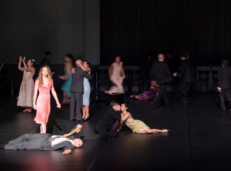 Scena ze spektaklu teatru tańca. Mężczyźni i kobiety przyjmują różne pozy: lezą na ziemi, chodzą, biegają, klaszczą, obejmują się. Część postaci jest rozmazana.