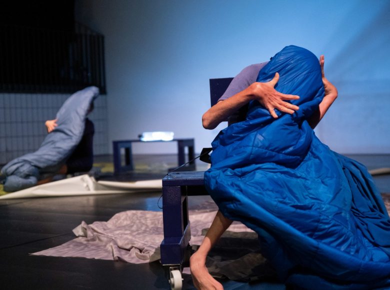 Scena. Na pierwszym planie na małym stoliku siedzi postać owinięta w niebieski śpiwór. Ma nienaturalnie wygięte kończyny, lekko się pochyla. W tle sceny widać drugą postać, która siedzi na krześle i też jest owinięta śpiworem.