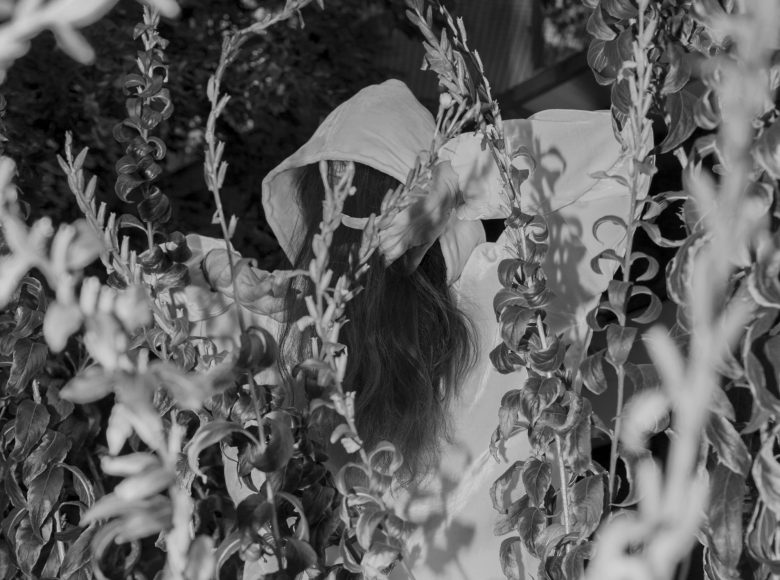 Czarno-biała fotografia. Kobieta ubrana w jasny strój z kapturem na głowie stoi pośród wysokich zarośli i traw. Ręce ma uniesione do góry, jej twarz zasłaniają długie rozpuszczone włosy.