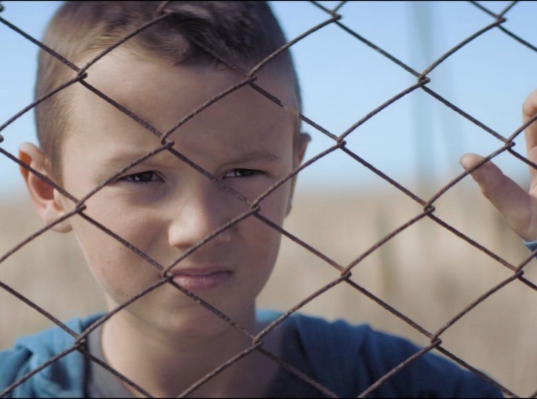 [ Kadr z filmu. Zbliżenie na twarz chłopca, który znajduje się za siatką ogrodzeniową. Ubrany w niebieską bluzę, patrzy przed siebie w skupieniu, dłonie ma trzyma na drutach siatki.