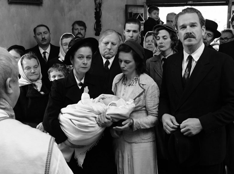 [ Czarno-biały kadr z filmu. W ciasnym wejściu kościoła tłoczy się kilkanaście osób w różnym wieku. Ubrani w stylu retro. Na pierwszym planie w rogu plecy księdza w szacie liturgicznej. W pierwszym rzędzie grupy stoją dwie kobiety i mężczyzna. Jedna z kobiet trzyma zawinięte w biały kocyk niemowlę i patrzy na księdza. Mężczyzna w garniturze patrzy w obiektyw. ]