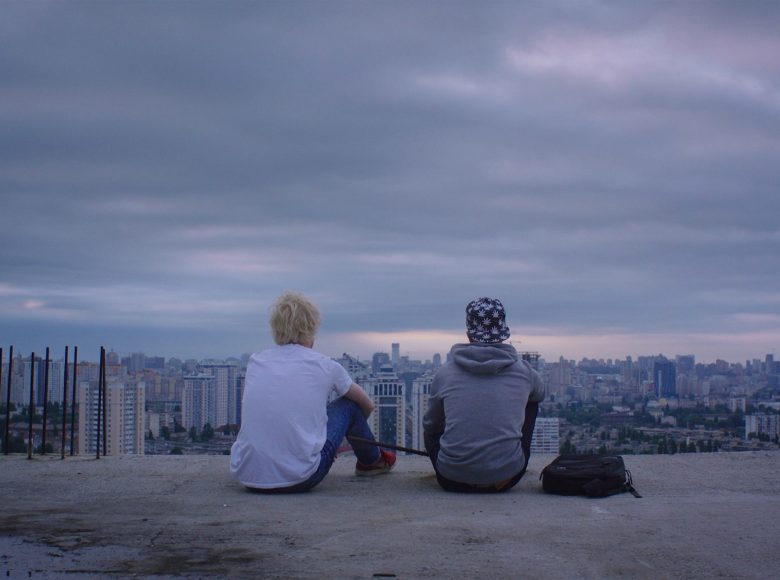 [ Kadr z filmu. Dwóch chłopaków siedzi tyłem do nas na dachu betonowej, nieskończonej budowli. Przed nimi rozciąga się panorama blokowisk dużego miasta. ]
