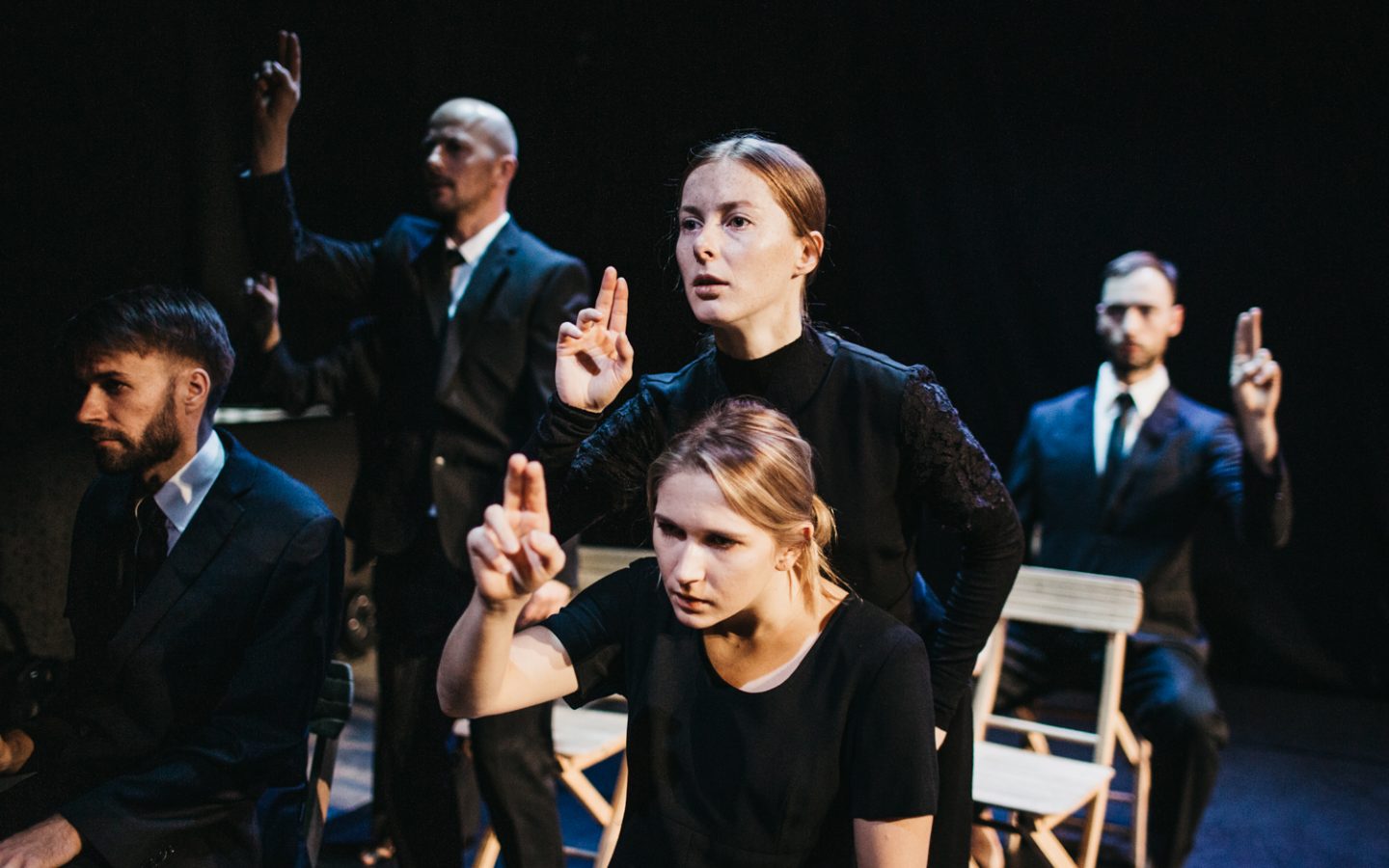 Zdjęcie. Grupa ludzi stoi lub siedzi na drewnianych krzesłach. Są ubrani na czarno. Wszyscy wykonują ten sam gest. Unoszą przed siebie jedną rękę i wystawiają dwa palce.