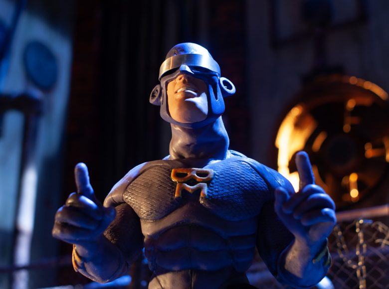 Kadr z filmu animowanego. Lalkowa postać superbohatera. Na piersi ma litery BM, na oczach mettalową przepaskę.