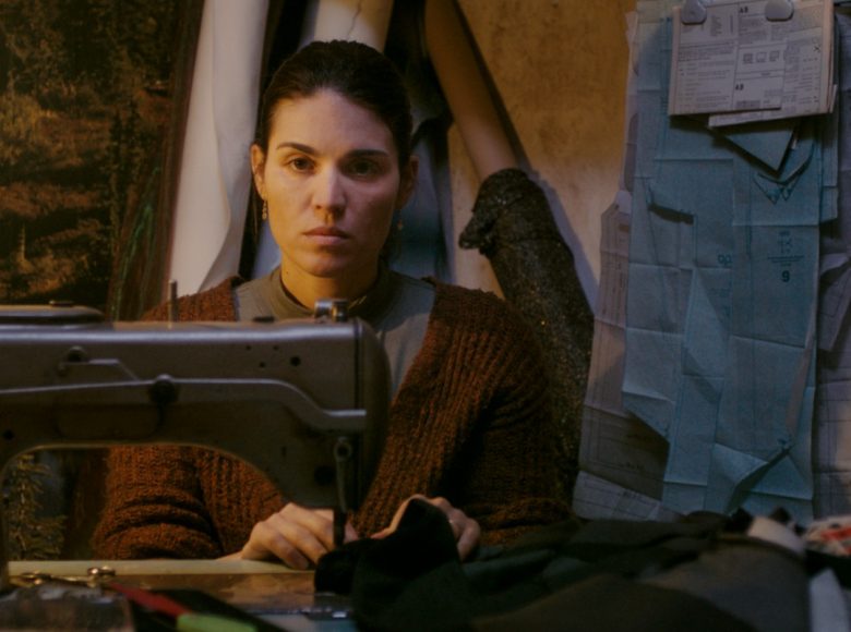 [ Kadr z filmu. Ciemne wnętrze. Za maszyną do szycia oświetloną gołą żarówką siedzi młoda kobieta. W dłoniach trzyma zszywaną tkaninę, ale spogląda na coś lub kogoś przed sobą. ]