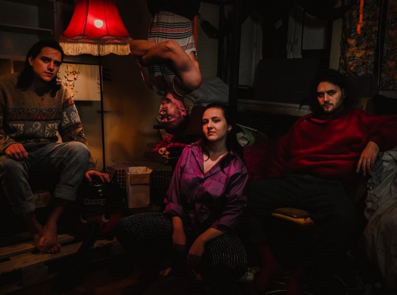 Zdjęcie. Grupa ludzi znajduje się w ciemnym pokoju. Pomieszczenie oświetla lampa z czerwonym abażurem. Kobieta i dóch mężczyzn siedzi na krześle. Trzeci mężczyzna zwisa do góry nogami z sufitu.