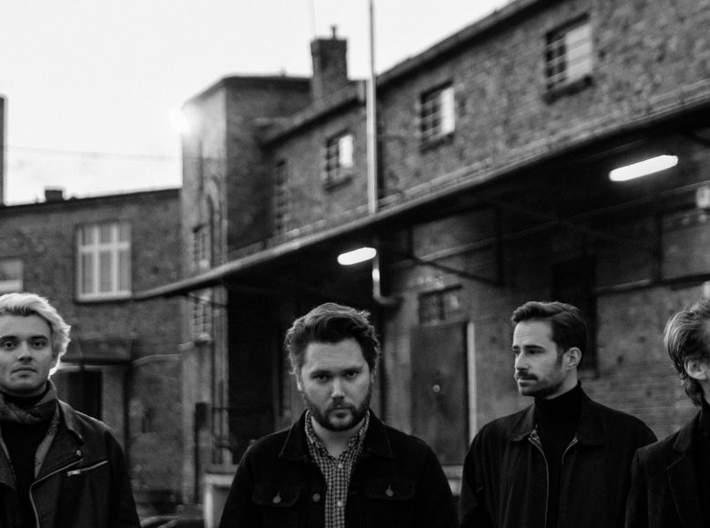 Czarno-białe zdjęcie. Czterech mężczyzn znajduje się na ulicy. Za nimi znajdują się ceglane budynki w stylu industrialnym.