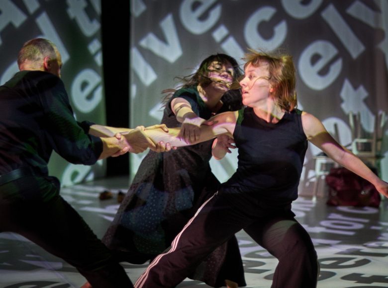Zdjęcie. Scena ze spektaklu teatru tańca "W moim początku jest mój kres". Na środku sceny stoi kobieta. Druga kobieta i mężczyzna szarpią ja za ręce. Na podłodze i ścianach odbijają się projekcje liter.