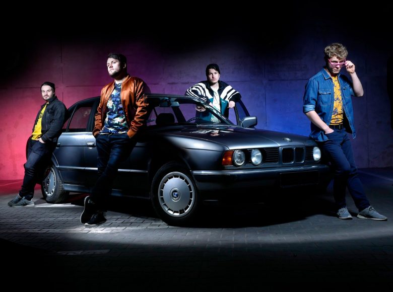 Zdjęcie. Czterech mężczyzn, w luźnych strojach stoi wokoło srebrnego BMW. Za nimi betonowa ściana, na którą pada czerwone i fioletowe światło.