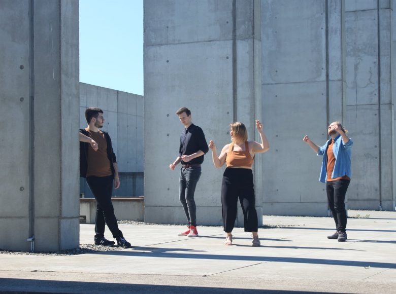 Zdjęcie. Trzech mężczyzn i jedna kobieta znajdują się na zewnątrz. Otaczają ich betonowe ściany. Jeden mężczyzna opiera się o betonowy mór. Pozostali tańczą.