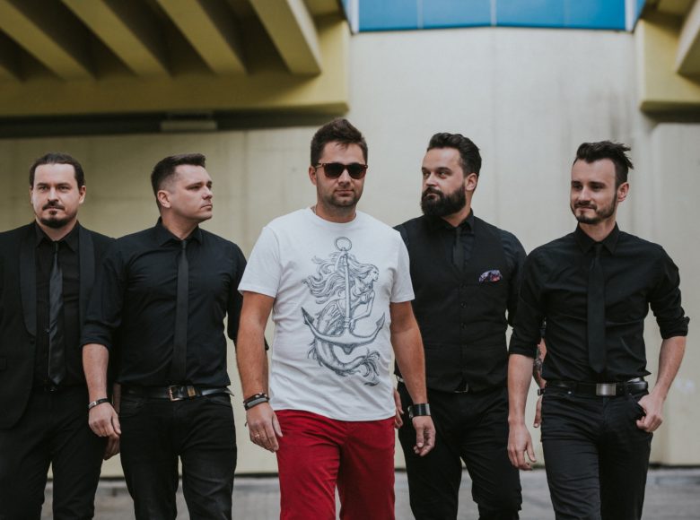 Zdjęcie. Czterech mężczyzn w czarnych koszulach i krawatach idzie przed siebie. Piąty mężczyzna na na sobie białą koszulkę z nadrukiem syreny z kotwicą i czerwone spodnie.