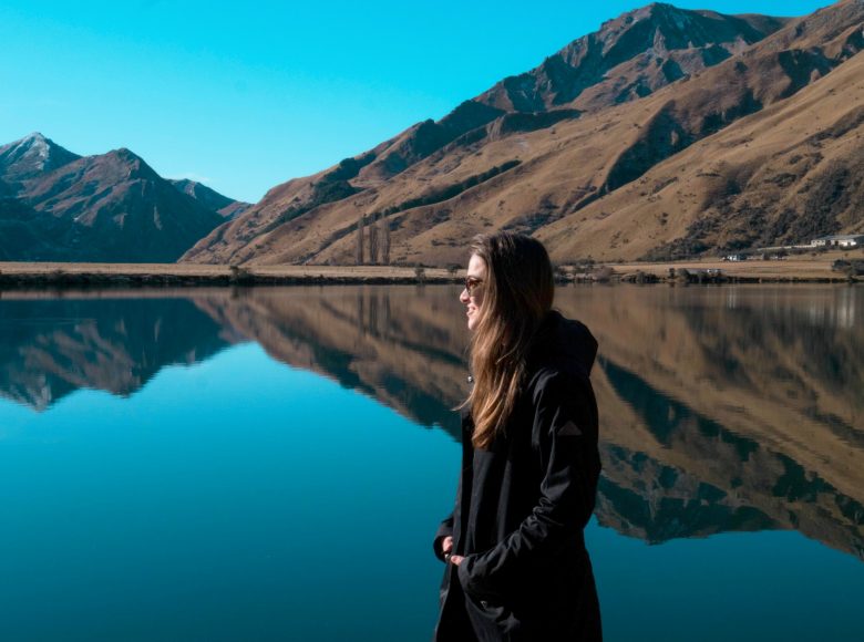 Zdjęcie w lekko pochmurny dzień. Na środku stoi bokiem do nas młoda dziewczyna z długim włosami. Jest ubrana w ciemny płaszczyk. W tle jezioro o niebiesko-zielonej wodzie. Po drugiej stronie jeziora zbocza gór porośniętych buro-zieloną roślinnością. Nad wszystkim niebieskie niebo.