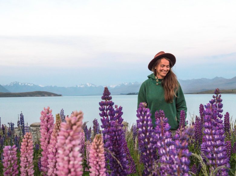 Na pierwszym planie fioletowe i różowe kwiaty zebrane w wysokie podłużne kwiatostany. Nieco z boku stoi uśmiechnięta dziewczyna z długimi brązowymi włosami. Jest ubrana w zieloną bluzę z kapturem i brązowy kapelusz w stylu kowbojskim. W tle wody jeziora. Bardzo daleko, po drugiej stronie jeziora widać góry z ośnieżonymi szczytami.
