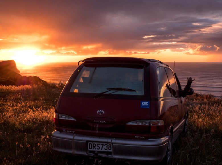 Zdjęcie o zachodzie słońce. Na skraju wybrzeża porośniętego trawami stoi spory samochód. W tle widać wodę oceanu, której kolory zmieniają się od granatu do złota. Po lewej stronie intensywny blask zachodzącego za horyzont słońca. Słońce odbija się w chmurach nadając im złoty kolor. Im dalej tym niebo jest ciemniejsze.