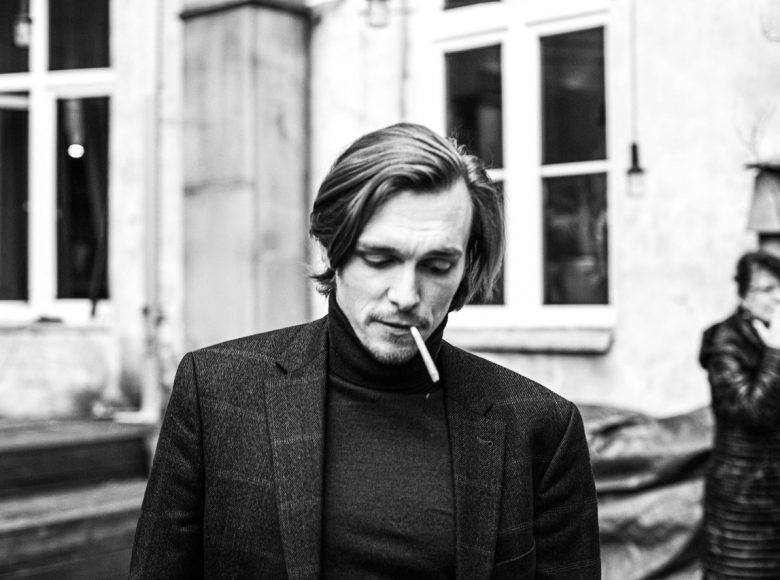 Czarno-białe zdjęcie. Młody mężczyzna z papierosem w ustach, widziany od torsu w górę. Na twarz opada mu lekko fala jasnych włosów. W tle wysokie okna budynku.