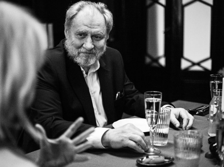 Mężczyzna z brodą siedzi za zastawionym stołem. Patrzy na osobę obok, widoczną częściowo, lekko się uśmiecha. Przed nim stoją szklanki i kieliszki.