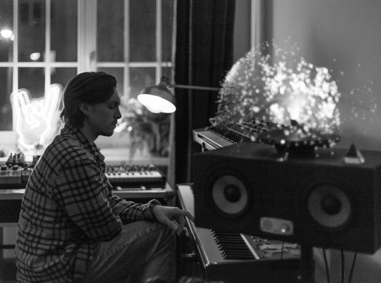 Czarno-białe zdjęcie. Młody mężczyzna widziany z profilu, siedzi przed dwupoziomowymi instrumentami klawiszowymi. Na pierwszym planie nieduży głośnik, na nim ozdobna lampa. W tle okno, neon dłoni i kolejne instrumenty.