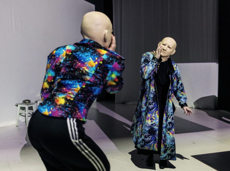 Zdjęcie. Scena ze spektaklu "W moim początku jest mój kres". Dwie osoby w kolorowych marynarkach i maskach przedstawiających twarz starego, łysego mężczyzny, stoi na przeciwko siebie i powtarza swoje ruchy jak w lustrzanym odbiciu.