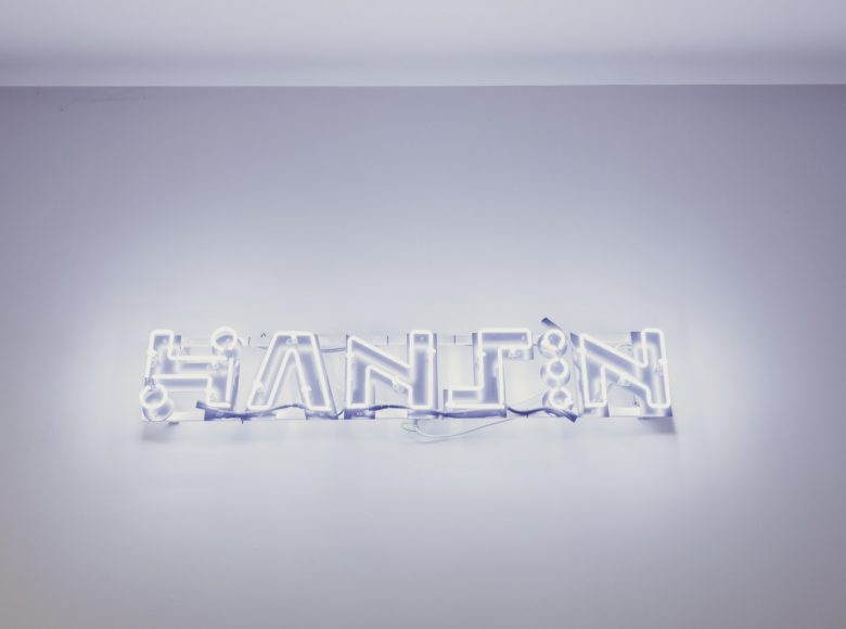 Zdjęcie przedstawia białą ścianę wewnątrz budynku rozświetloną wiszącym na niej neonem, który układa się w napis Hansen.