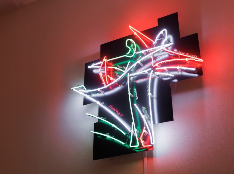 Zdjęcie przedstawia jasną ścianę wewnątrz budynku na której wisi kolorowy neon przedstawiający łyżwiarzy.