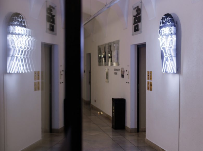 Zdjęcie przedstawia korytarz wewnątrz budynku. Na ścianie obok drzwi prowadzących do pomieszczenia wisi jasno świecący neon przedstawiający chłopięcą postać żonglującą piłeczkami. Piłeczki jednocześnie stanowią literę "o". Nad głową postać ma napis "tak". Neon migając układa się w napis "o tak o".