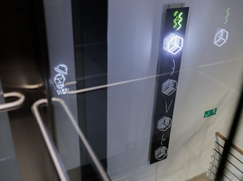 Zdjęcie przedstawia widok ze szklanej windy wewnątrz budynku. Lekko w dole widać fragment klatki schodowej i półpiętro z białą ścianą. Na ścianie wisi podłużny neon przedstawiający spadające i migające kostki cukru.