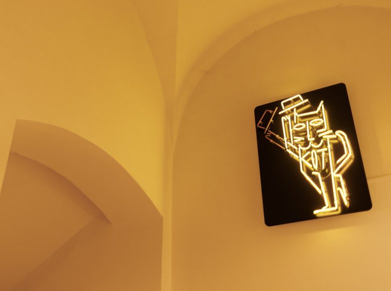 Zdjęcie przedstawia jasne ściany i sufit na korytarzu wewnątrz budynku. Na jednej ścianie na czarnym tle wisi jasno świecący neon przedstawiający sylwetkę stojącego kota, który jedną z łap zdejmuje z głowy kapelusz.