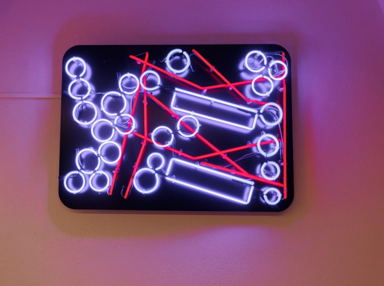 Zdjęcie przedstawia jasną ścianę wewnątrz budynku. Na ścianie wisi neon na czarnym tle, świeci na fioletowo i czerwono. Świecące linie, koła i prostokąty układają się w plan jednego z lubelskich osiedli.