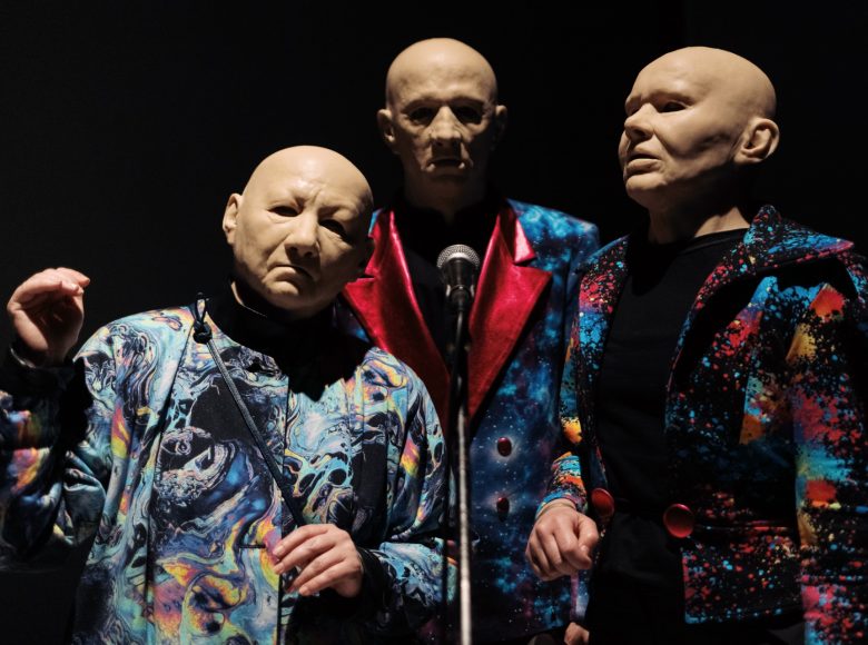 Zdjęcie. Scena ze spektaklu "W moim początku jest mój kres". Przedstawia trzy osoby w identycznych maskach, przedstawiających twarz starego, łysego mężczyzny. Stoją przed mikrofonem. Tło ciemne, jednolite.