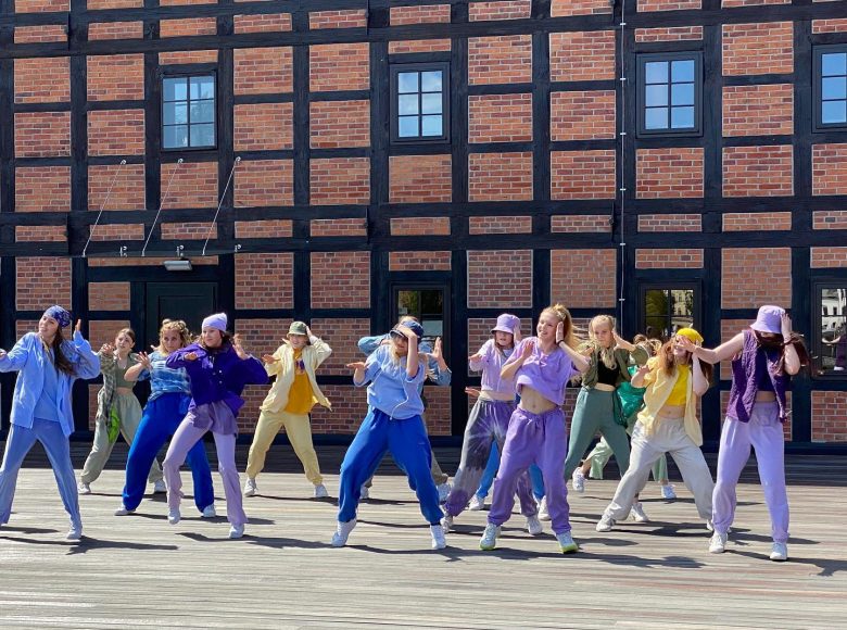 Zdjęcie. Grupa ludzi w kolorowych dresach tańczy na drewnianym parkiecie. Za nimi stoi ceglany budynek.