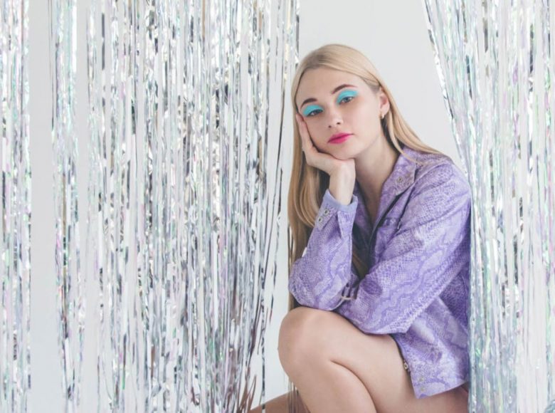 Zdjęcie. Młoda dziewczyna w jaskrawym makijażu i fioletowej kurtce imitującej wężową skórę siedzi pomiędzy srebrnymi zasłonami.