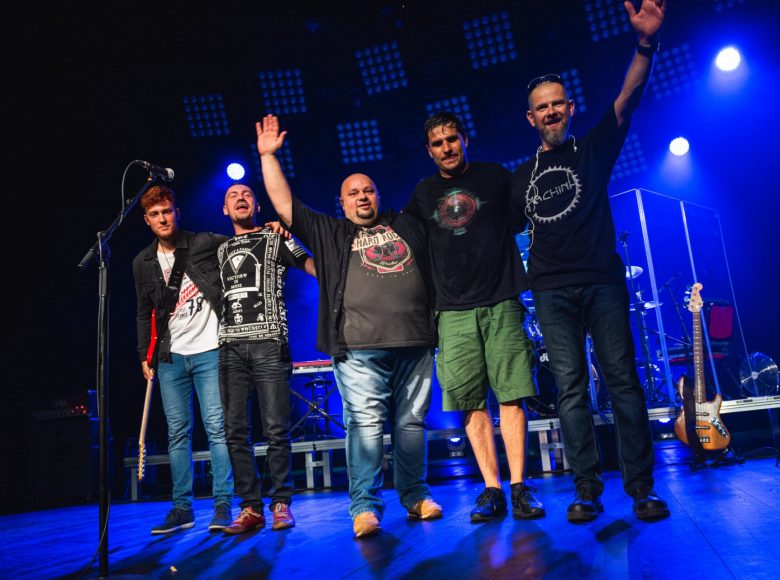 Zdjęcie. Grupa mężczyzn stoi na scenie. Obejmują się za ramiona, pozują do zdjęcia. Za nimi znajduje się platforma z instrumentami. Na scenę pada niebieskie światło.