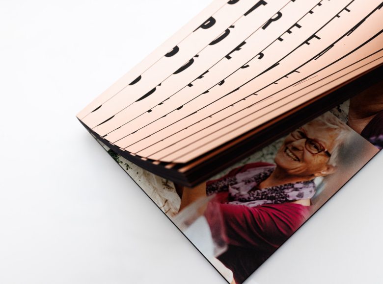 Zdjęcie uchylonej książki. Okładka w kolorze kremowym, przecinają ją czarne, pionowe pasy i kawałki liter układające się w napis Strefa Półcienia. Na widocznej stronie zdjęcie starszej kobiety, z krótkimi siwymi włosami, fioletowej apaszce w kwiaty i bordowej bluzce. Kobieta siedzi przy stole, jest uśmiechnięta.